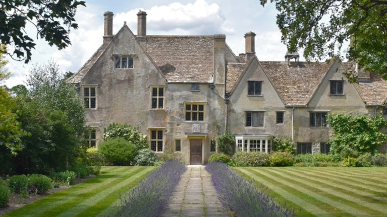 A landscape shot of the East Garden in Avebury Manor Garden showing flower beds on the left and a cut lawn on the right. Purple lavender lines both sides of the footpath through the middle of the photo, with the path leading to the door of the 16th Century Avebury Manor house.